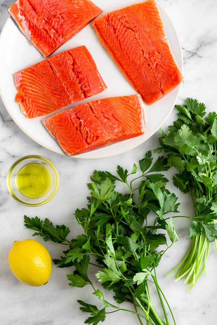 An overhead shot with a plate of salmon filets, fresh parsley, a lemon, and a bowl of olive oil.