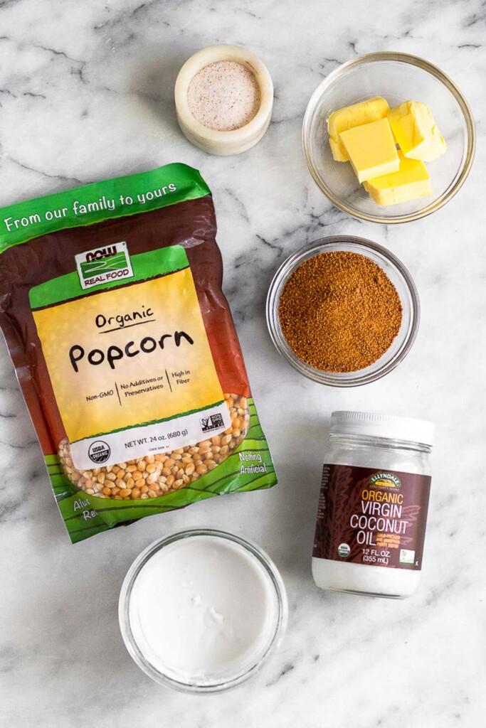 White marble counter with a dish of salt, a bowl of slices of butter, a bowl of coconut sugar, a jar of coconut oil, a jar of coconut milk, and bag of popcorn kernels.