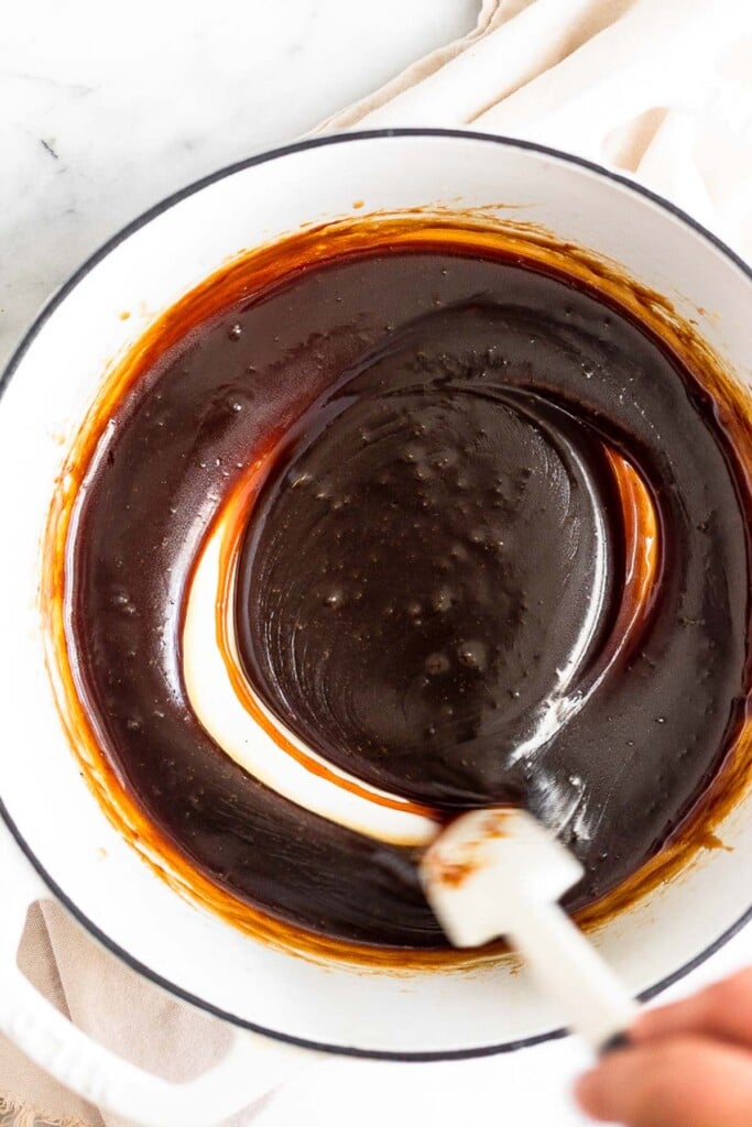 Large white dutch oven with homemade caramel sauce in it being stirring with a spatula.