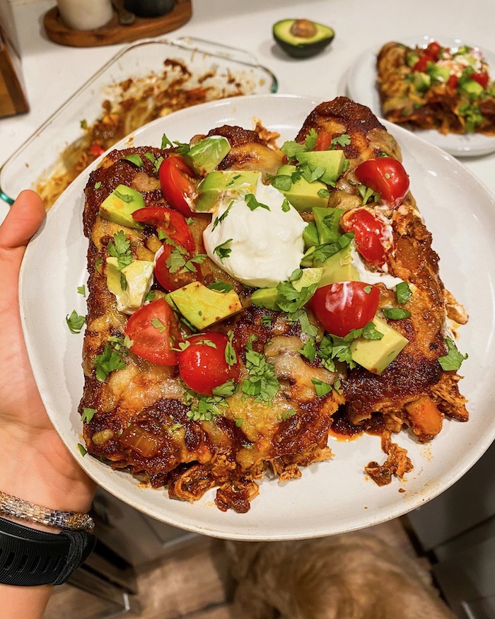 Shot of chicken enchiladas on a plate topped with avocado, tomatoes, sour cream, and herbs.