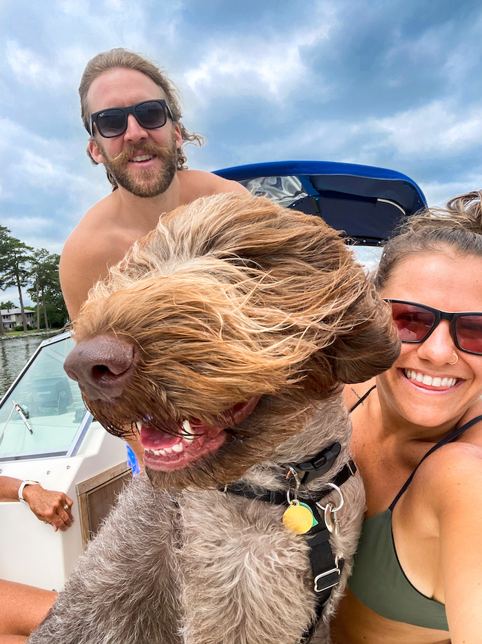 Woman and man in sunglasses with a wirehaired pointing griffon dog in-between them on a boat.