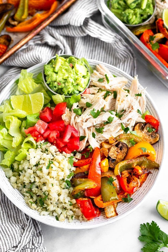 Chicken burrito bowl with shredded chicken, cauliflower rice, fajita veggies, lettuce, tomato, guacamole, and a lime wedge.