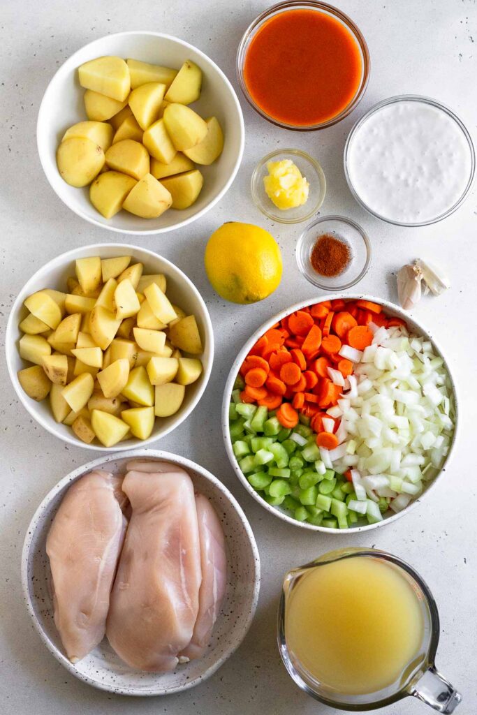 Overhead shot of a bowl of hot sauce, a bowl of coconut milk, a bowl of ghee, a bowl of spices, 2 garlic cloves, a bowl of diced carrots, celery, and onions, a cup of broth, a plate of raw chicken breasts, a bowl of diced potatoes, and a bowl of quartered potatoes.