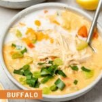 A bowl of buffalo chicken soup topped with coconut milk and green onions and a spoon in it. Behind it is another bowl and half a lemon.