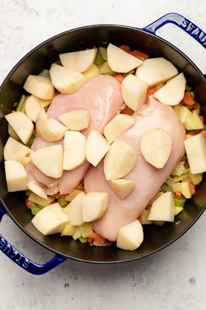 Overhead shot of a large pot with cooked veggies on the bottom, two raw chicken breasts, and quartered potatoes on top.
