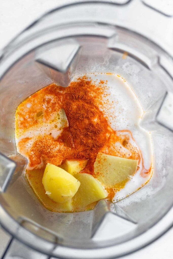 Blender with cooked potatoes, coconut milk, hot sauce, and spices in it.