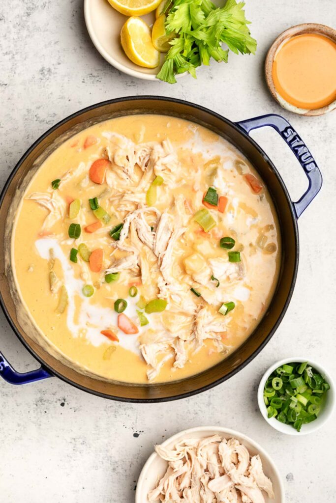 Large pot of buffalo chicken soup garnished with coconut cream and cream onions. Around it is a bowl of shredded chicken, a bowl of green onions, a bowl of buffalo sauce, and a plate of lemons and celery.