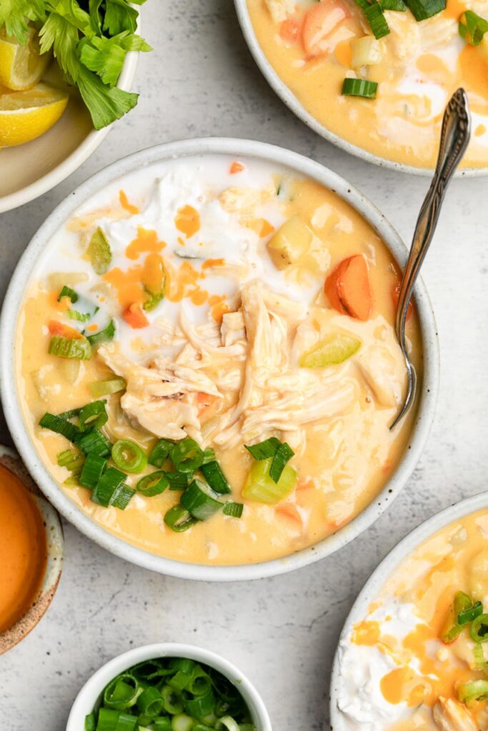 A bowl of creamy buffalo chicken soup topped with green onions and coconut milk. Around it is two more bowls of soup, a bowl of green onions, a bowl of hot sauce, and a bowl of lemons.