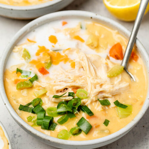 A bowl of buffalo chicken soup topped with coconut milk and green onions and a spoon in it. Behind it is another bowl and half a lemon.