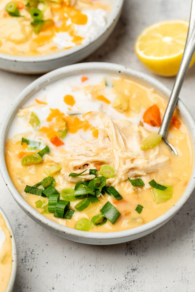 A bowl of buffalo chicken soup topped with coconut milk and green onions and a spoon in it. Behind it is another bowl and half a lemon.