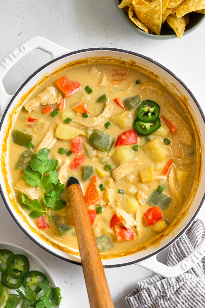Large stockpot filled with creamy chicken potato soup garnished with jalapeño and cilantro. A ladle is coming out of it. Around it is a plate of sliced jalapeños, a towel, and a bowl of chips.