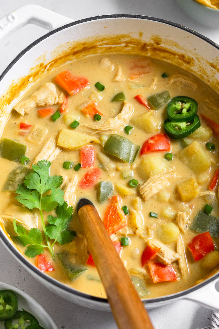 Large dutch oven filled with healthy creamy chicken soup topped with cilantro, green onions, and jalapeños with a ladle coming out of it.