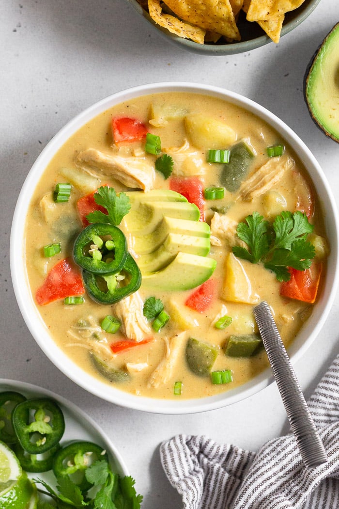 Overhead shot of cheesy chicken soup in a bowl topped with avocado, jalapeños, and herbs. A spoon is in the bowl and around it is a plate with more herbs, an avocado, a bowl of chips, and a kitchen towel.