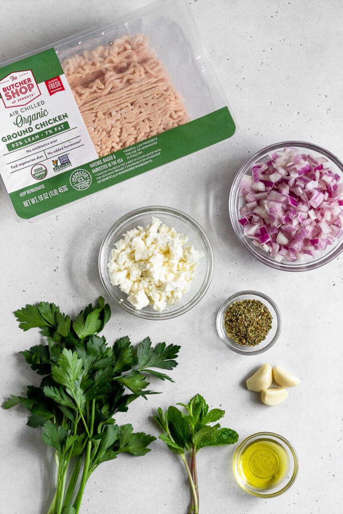 White concrete counter with a package of raw ground chicken, a bowl of diced red onions, a small bowl of dried herbs, 3 garlic cloves, a small bowl of olive oil, a few sprigs of mint, a bunch of fresh parsley, and a bowl of crumbled feta.