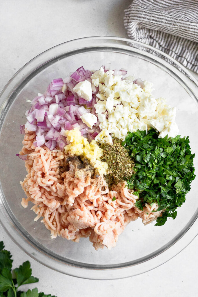 Large glass mixing bowl of ground chicken, diced onion, minced garlic, crumbled feta, chopped herbs, and spices before it is mixed together. Next to the bowl is a striped towel and some fresh herbs.
