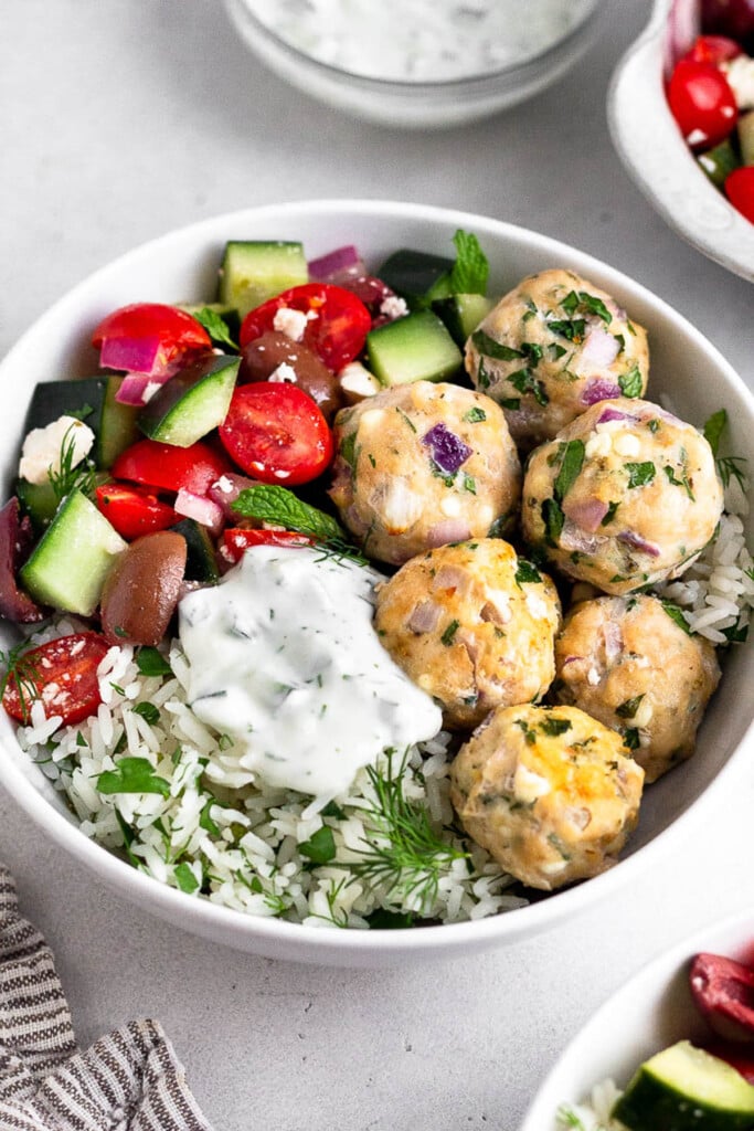 Chicken feta meatballs with white rice mixed with herbs, tzatziki sauce, and a tomato cucumber salad in a white bowl. It is garnished with fresh herbs. Around the bowl is a striped kitchen towel, a bowl of tzatziki sauce, and two bowls of tomato cucumber salad.