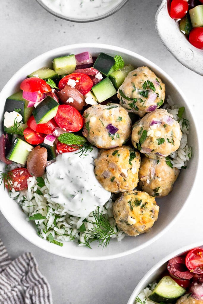 Mediterranean meatballs in a bowl with herby rice, greek salad with tomatoes, cucumber, olives, and feta, and tzatziki sauce. Around the bowl is a bowl of tzatziki sauce, two bowls with greek salad, and a striped kitchen towel.