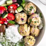 Greek chicken meatballs with herby rice, a greek salad with tomatoes, cucumbers, olives, and feta, and tzatziki sauce in a white bowl.