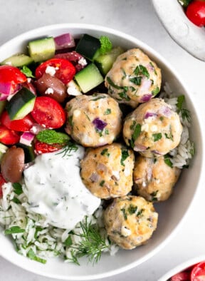 Greek chicken meatballs with herby rice, a greek salad with tomatoes, cucumbers, olives, and feta, and tzatziki sauce in a white bowl.