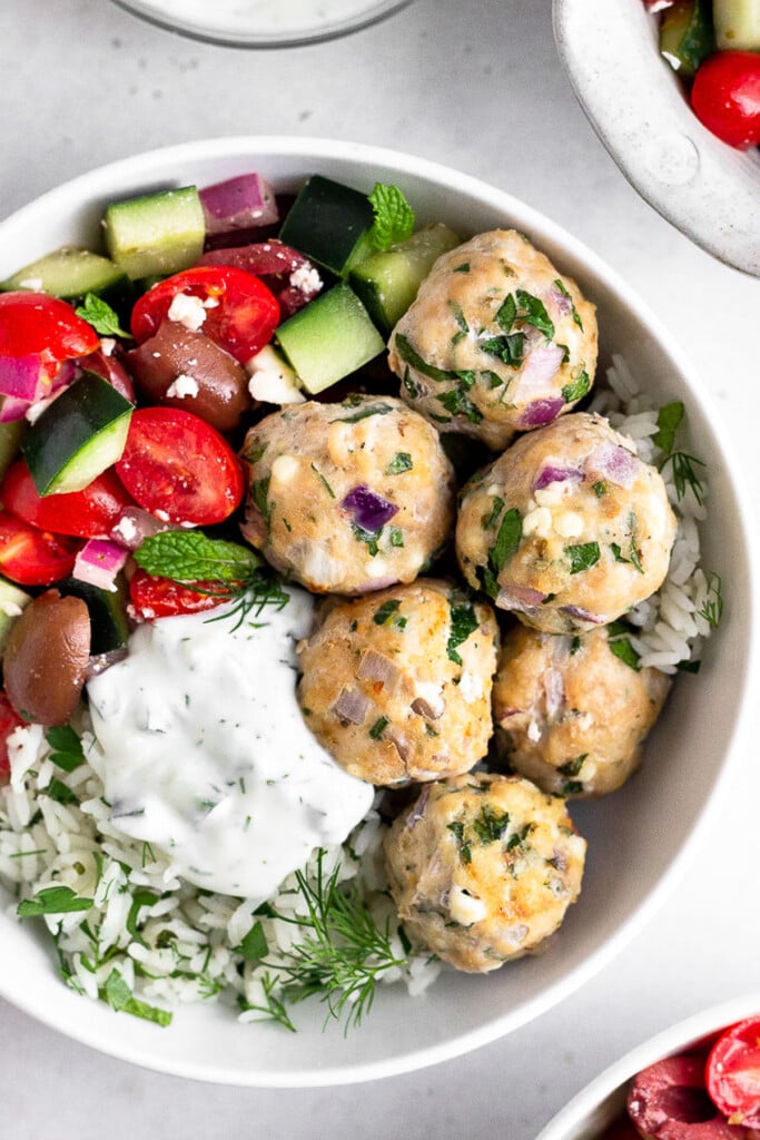 Greek chicken meatballs with herby rice, a greek salad with tomatoes, cucumbers, olives, and feta, and tzatziki sauce in a white bowl.