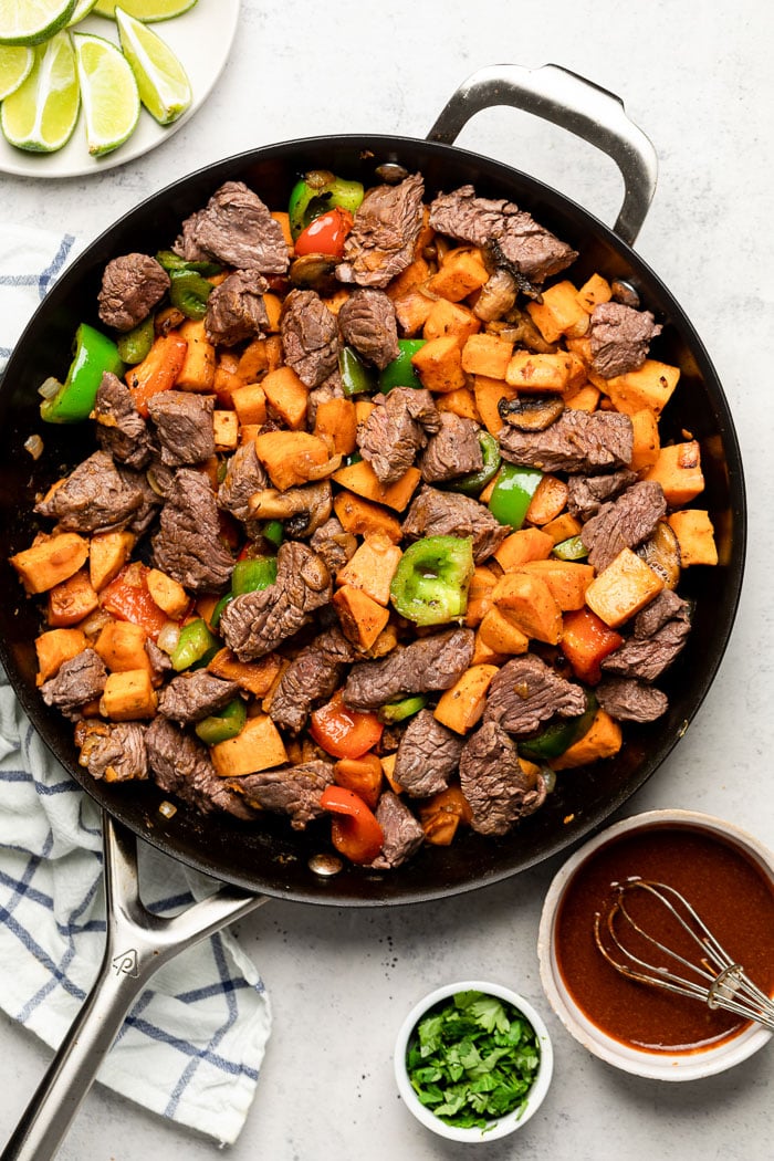 Steak bites with sweet potatoes, peppers, and mushrooms in a large pan. Next to it is a bowl of chipotle sauce, cilantro, and lime wedges.