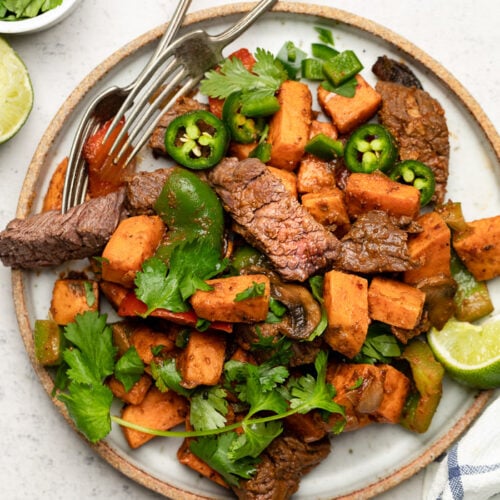 Plate with steak and sweet potato topped with cilantro and jalapeños. Next to it is a stripped towel, lime wedge, and a bowl of cilantro.