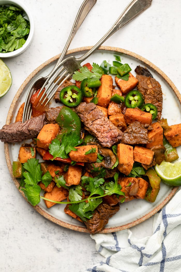 Plate with steak and sweet potato topped with cilantro and jalapeños. Next to it is a stripped towel, lime wedge, and a bowl of cilantro.
