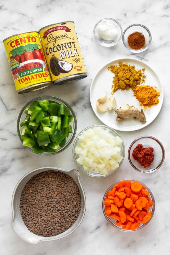 White marble counter with a bowl of coconut oil, a bowl of cayenne pepper, a plate of curry powder, ground turmeric, a nob of fresh ginger, and 3 cloves of garlic, a bowl of red curry paste, a bowl of diced onions, a bowl of sliced carrots, a bowl of uncooked brown lentils, a bowl of diced green bell peppers, a can of diced tomatoes, and a can of coconut milk.