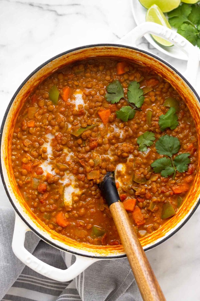 A large pot of lentil curry soup garnished with cilantro and coconut milk. A ladle is in the pot. Next to the pot is a plate of lime wedges and fresh cilantro.