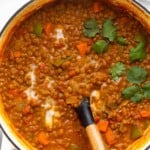 Curry lentil soup garnished with coconut milk and cilantro in a large white dutch oven with a spoon in it. Off to the side is a plate of lime wedges.