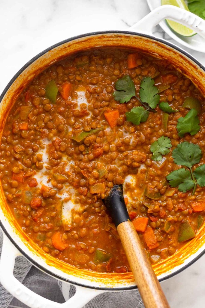 Curry lentil soup garnished with coconut milk and cilantro in a large white dutch oven with a spoon in it. Off to the side is a plate of lime wedges.