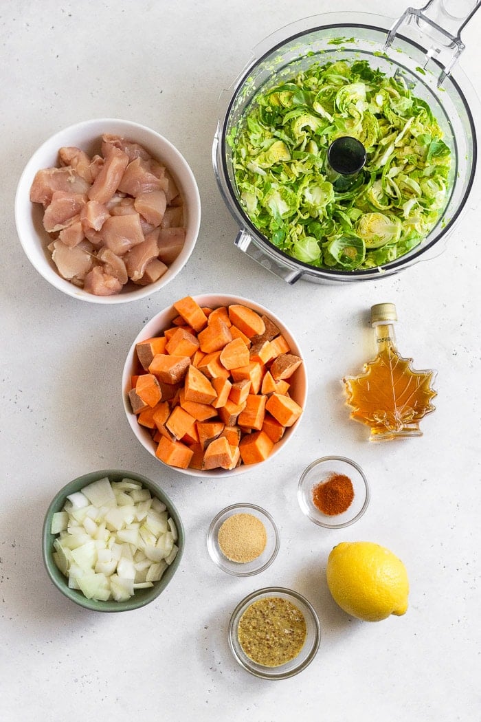 Overhead shot of a food processor filled with shredded Brussel sprouts, a jar of maple syrup, lemon, two small bowls of spices, a bowl of mustard, bowl of chopped onion, bowl of diced sweet potatoes, and a bowl of raw cubed chicken breasts.