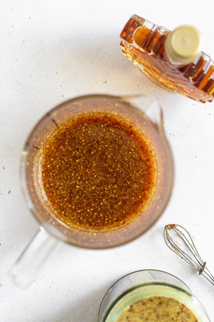 Maple mustard sauce in a mixing glass with a bottle of maple syrup, a small whisk, and jar of mustard next to it.