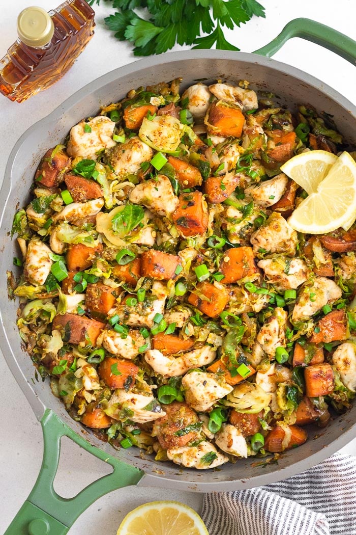 Maple mustard chicken recipe with sweet potatoes and Brussel sprouts in a pan. Around it is a jar of maple syrup, fresh parsley, and half a lemon.