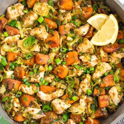 A pan of maple mustard chicken skillet with sweet potatoes and Brussels with lemon wedges on top.