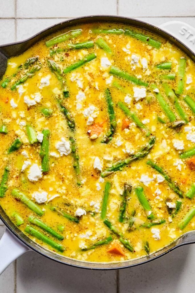 Large enameled skillet filled with a mixture of whisked eggs, goat cheese, and veggies before it is baked.