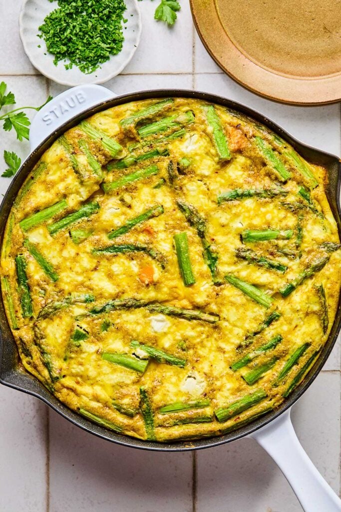 Asparagus goat cheese frittata with sweet potatoes in a large enameled skillet. Next to the skillet some fresh parsley, a small plate with chopped parsley, and an empty plate.