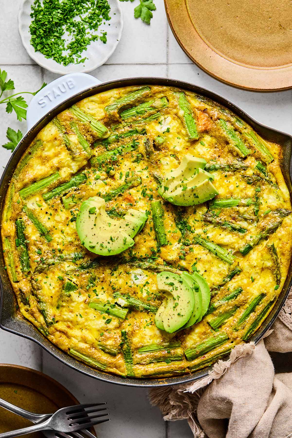 Asparagus frittata with sweet potatoes and goat cheese in a large enameled skillet. It is topped with sliced avocado and fresh parsley. Around the pan is a tan kitchen towel, a plate with two forks on it, a small dish of chopped fresh parsley, and another empty plate.