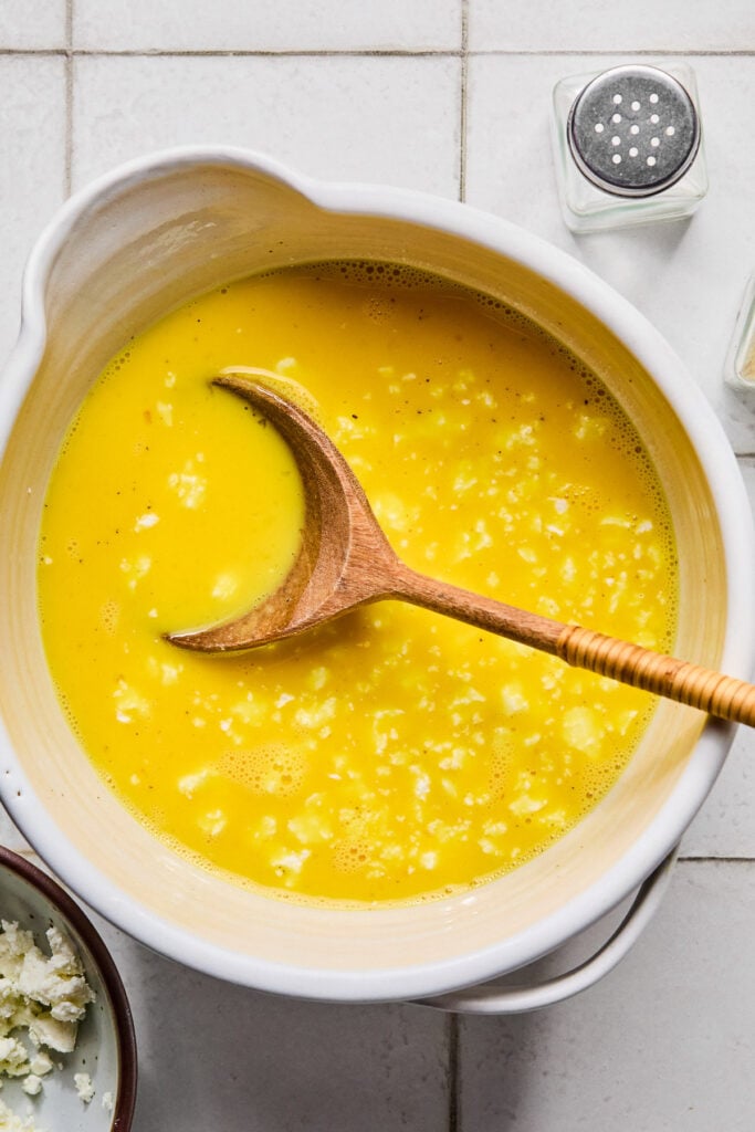 A large white mixing bowl of whisked eggs, milk, and goat cheese. A wooden spoon is also in the bowl. Next to the bowl is a salt shaker and a bowl of goat cheese.