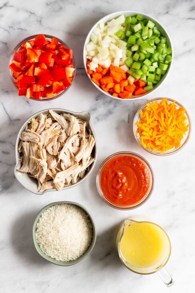 White marble counter with a bowl of diced onion, celery, and carrots, a bowl of shredded cheddar cheese, a bowl of buffalo sauce, a measuring cup of broth, a bowl of uncooked white rice, a bowl of shredded chicken, and a bowl of diced red bell peppers.