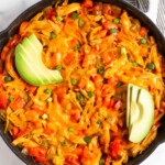 Buffalo chicken casserole topped with melted cheese in large cast iron skillet. It is garnished with sliced avocado and green onions. Next to it is a bowl of more green onions and half an avocado.