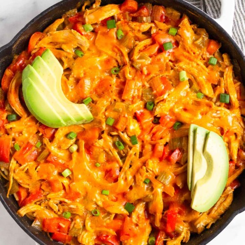 Buffalo chicken casserole topped with melted cheese in large cast iron skillet. It is garnished with sliced avocado and green onions. Next to it is a bowl of more green onions and half an avocado.