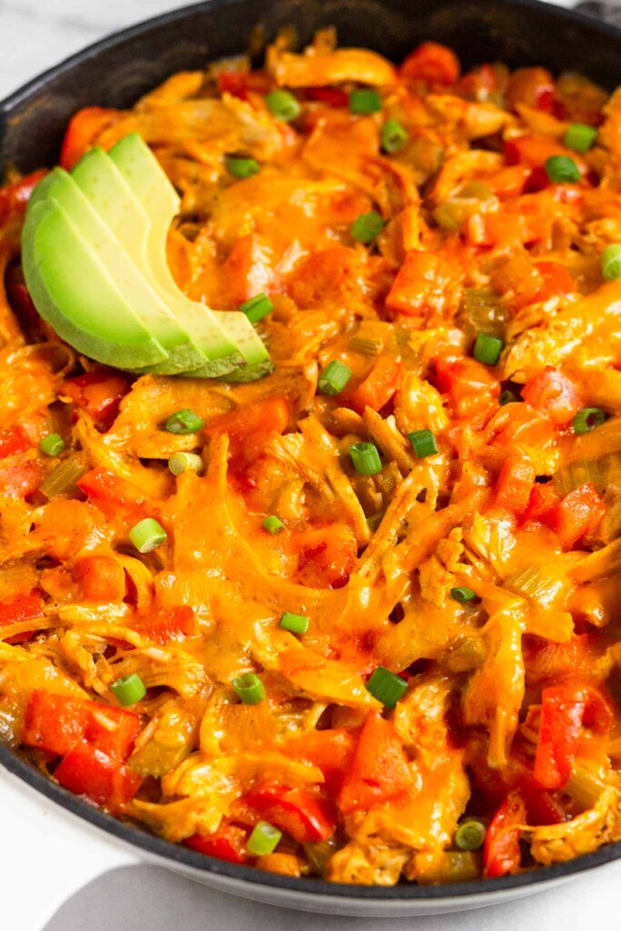 Buffalo chicken and rice casserole topped with melted cheese in a large cast iron skillet. It is garnished with sliced avocado and sliced green onions.