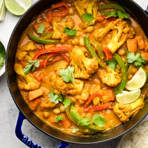 Large dutch oven filled with vegan cauliflower chickpea curry garnished with cilantro. Next to it is lime wedges and a bowl of white rice.