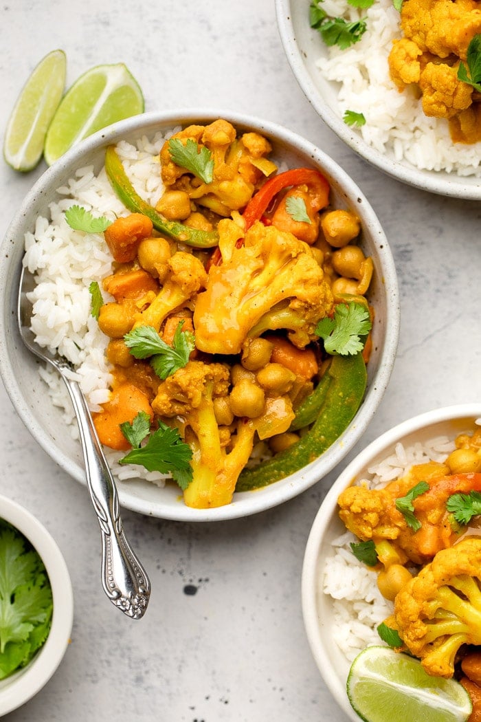 Vegan chickpea curry with cauliflower and veggies in a bowl with rice. It is garnished with cilantro. Next to it is more bowls of curry and some lime wedges.