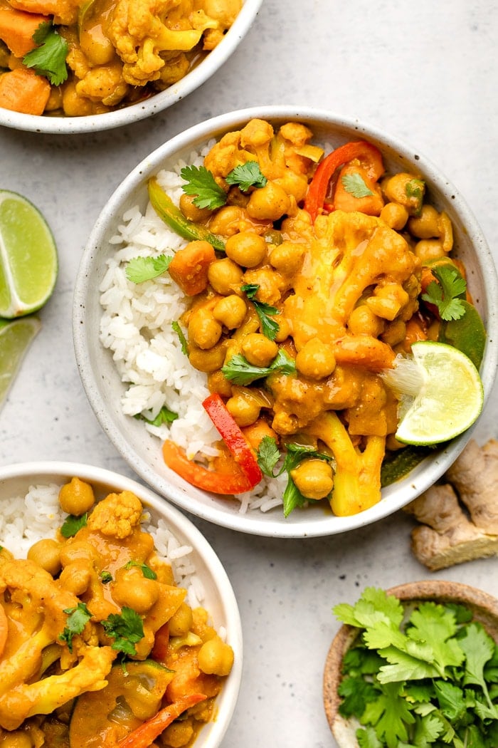 Vegan curry recipe in a bow with rice, a lime wedge, and cilantro. Next to is more bowls of curry, lime wedges, a small bowl of cilantro, and a knob of ginger.