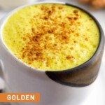 Close up of a mug filled with a golden milk latte sprinkled with cinnamon. Behind the mug is a piece of fresh turmeric, a piece of fresh ginger, and a jar of milk.