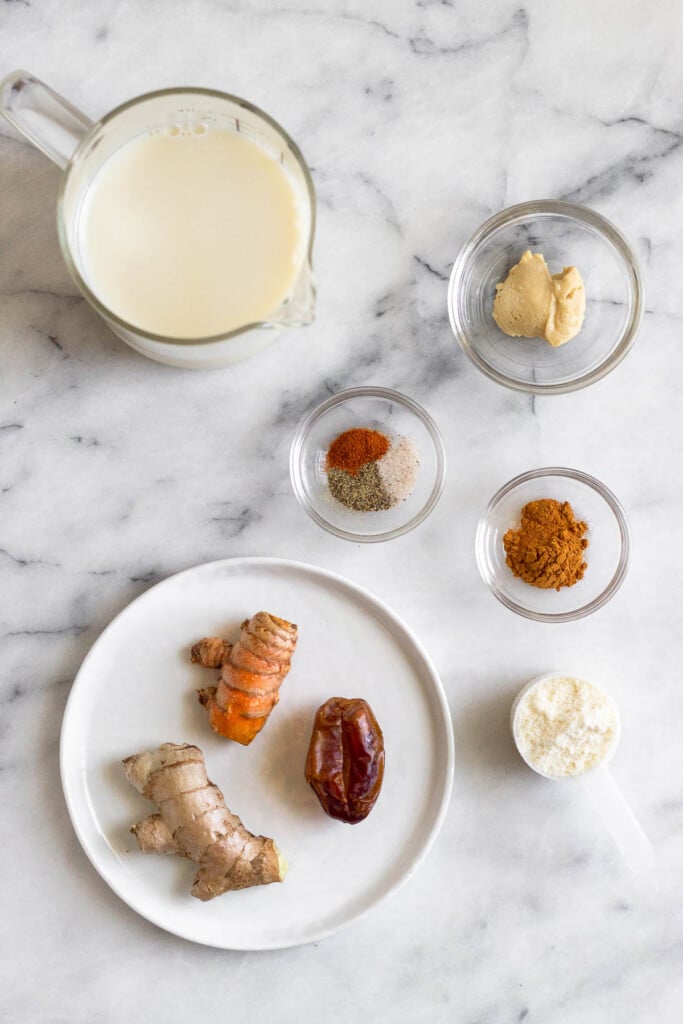 White marble counter with a small bowl of a cashew butter, a small bowl of spices, a small bowl of cinnamon, a scoop of collagen, a plate with a knob of fresh ginger, a piece of fresh turmeric, and a date, and a cup filled with milk.