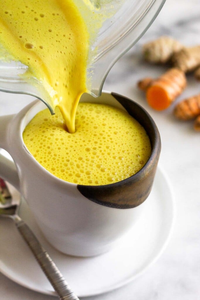 Turmeric latte being poured into a mug from a blender. The mug is sitting on a small plate with a spoon on it. Behind the mug are pieces of fresh turmeric and ginger.