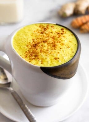 Golden milk latte sprinkled with cinnamon in a white mug sitting on a plate with a spoon on it. Behind it are pieces of fresh turmeric and ginger and a glass jar of milk.